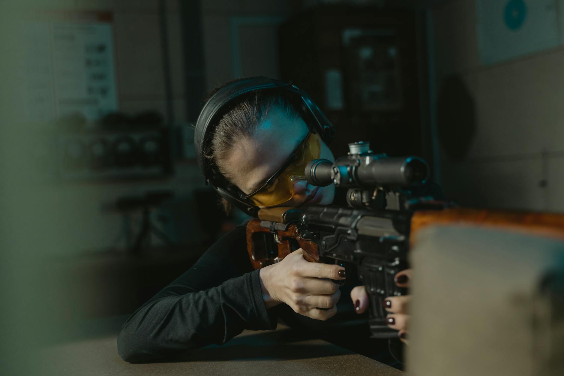 A woman with protective gear aiming a sniper rifle at an indoor shooting range.