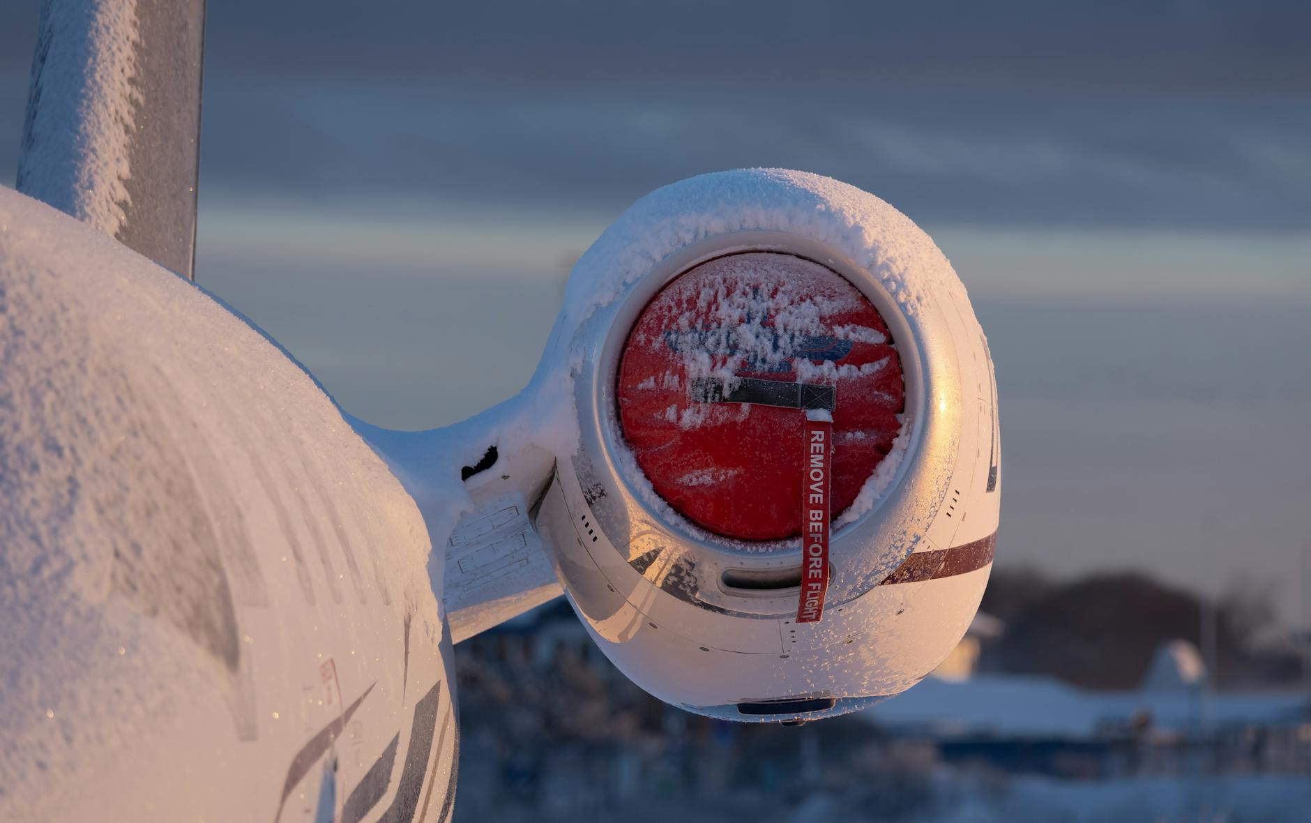 Airplane engine covered in snow at sunrise, showcasing winter aviation scene. Remove Before Flight tag visible.