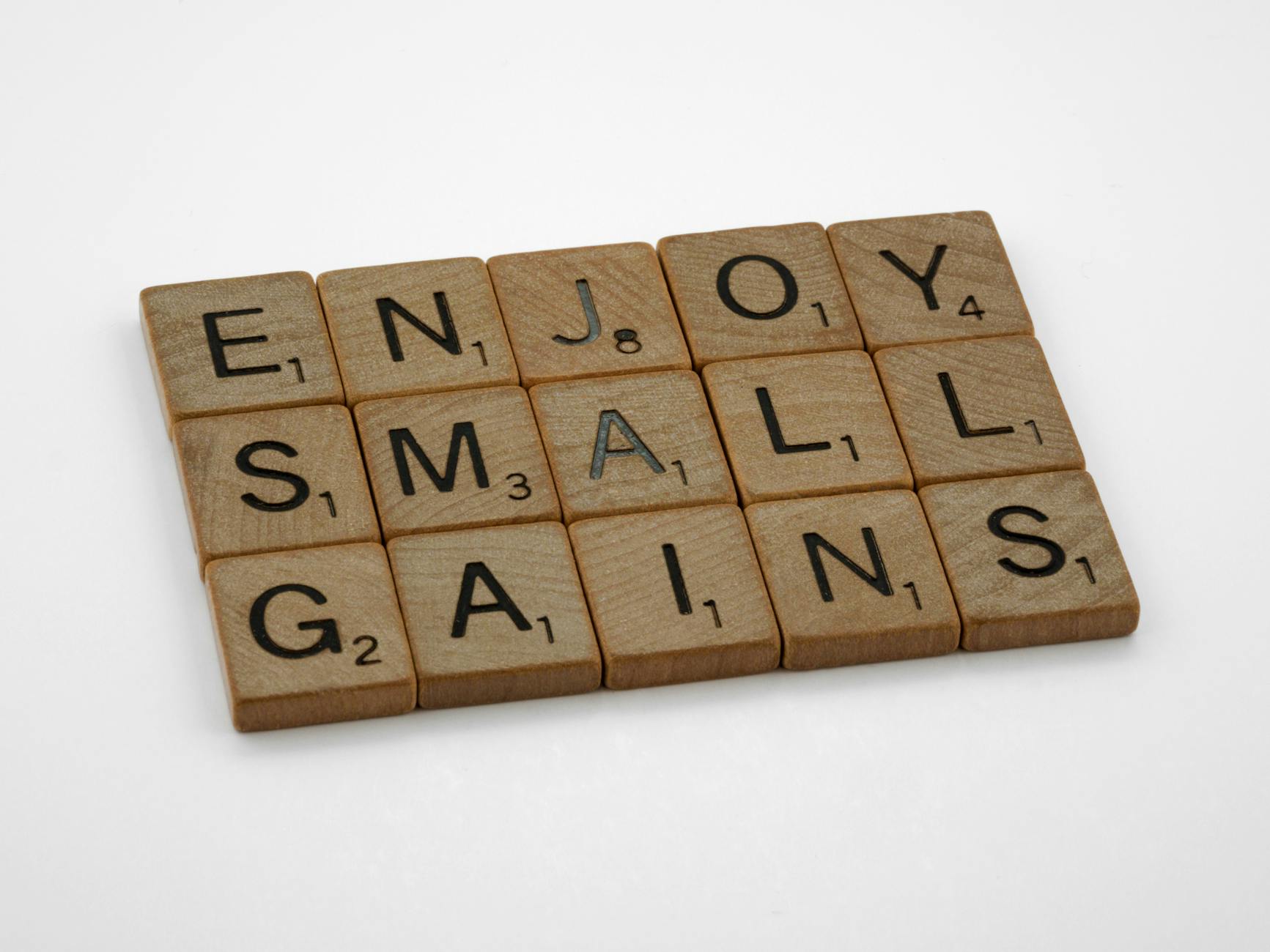 Close-up of Scrabble tiles spelling 'Enjoy Small Gains' on a white surface.