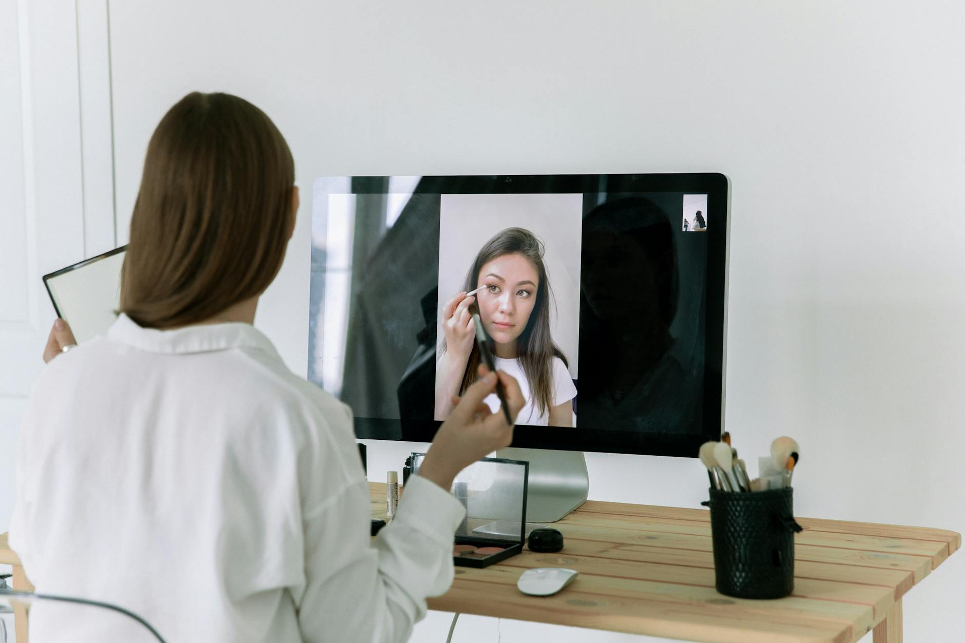 Woman following an online beauty tutorial, applying makeup using brushes in front of a computer screen.