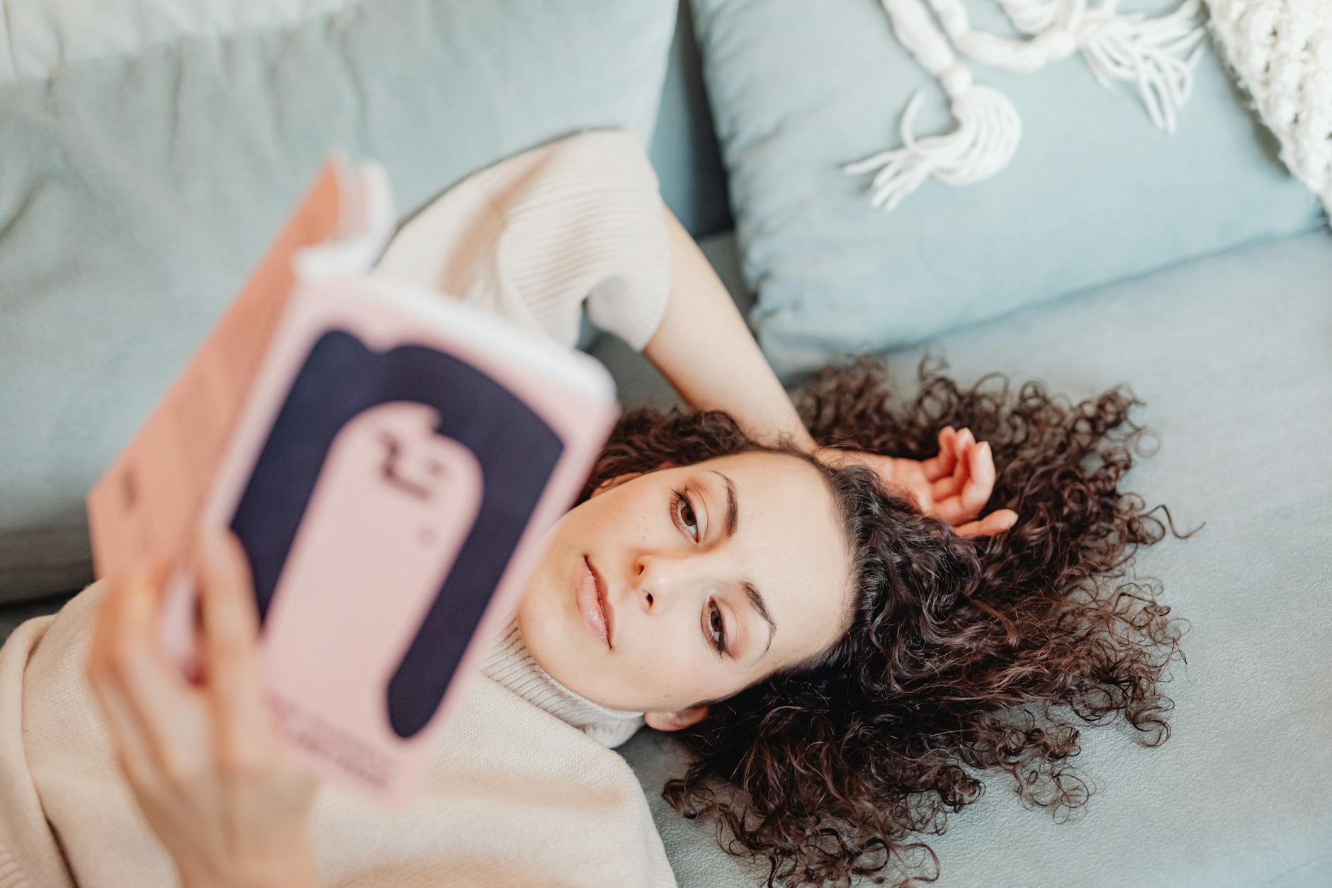 Woman with curly hair enjoying a peaceful reading session on a plush sofa, emphasizing relaxation and leisure.