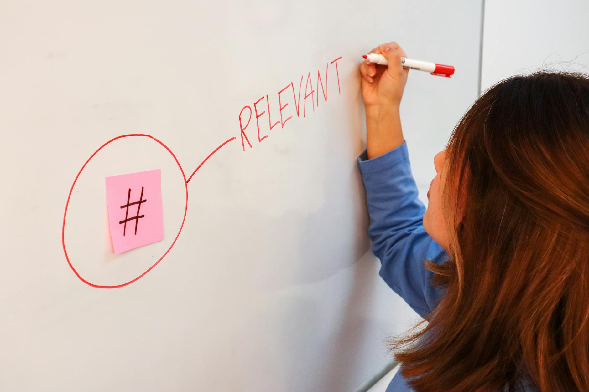 Woman writing the word 'RELEVANT' on a whiteboard with a red marker beside a sticky note hashtag.
