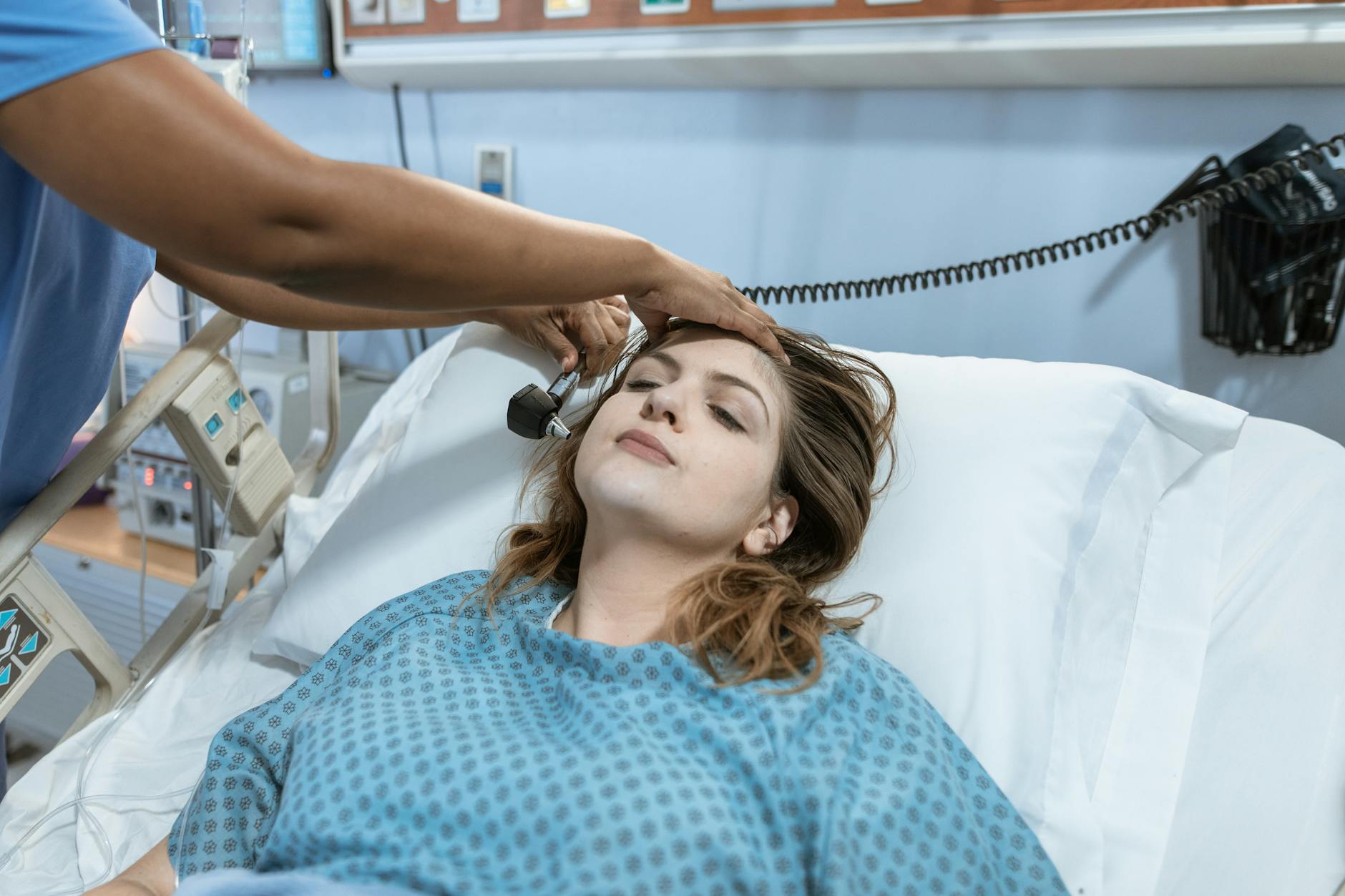 A female patient in a hospital bed undergoing a check-up with a healthcare worker using an otoscope.