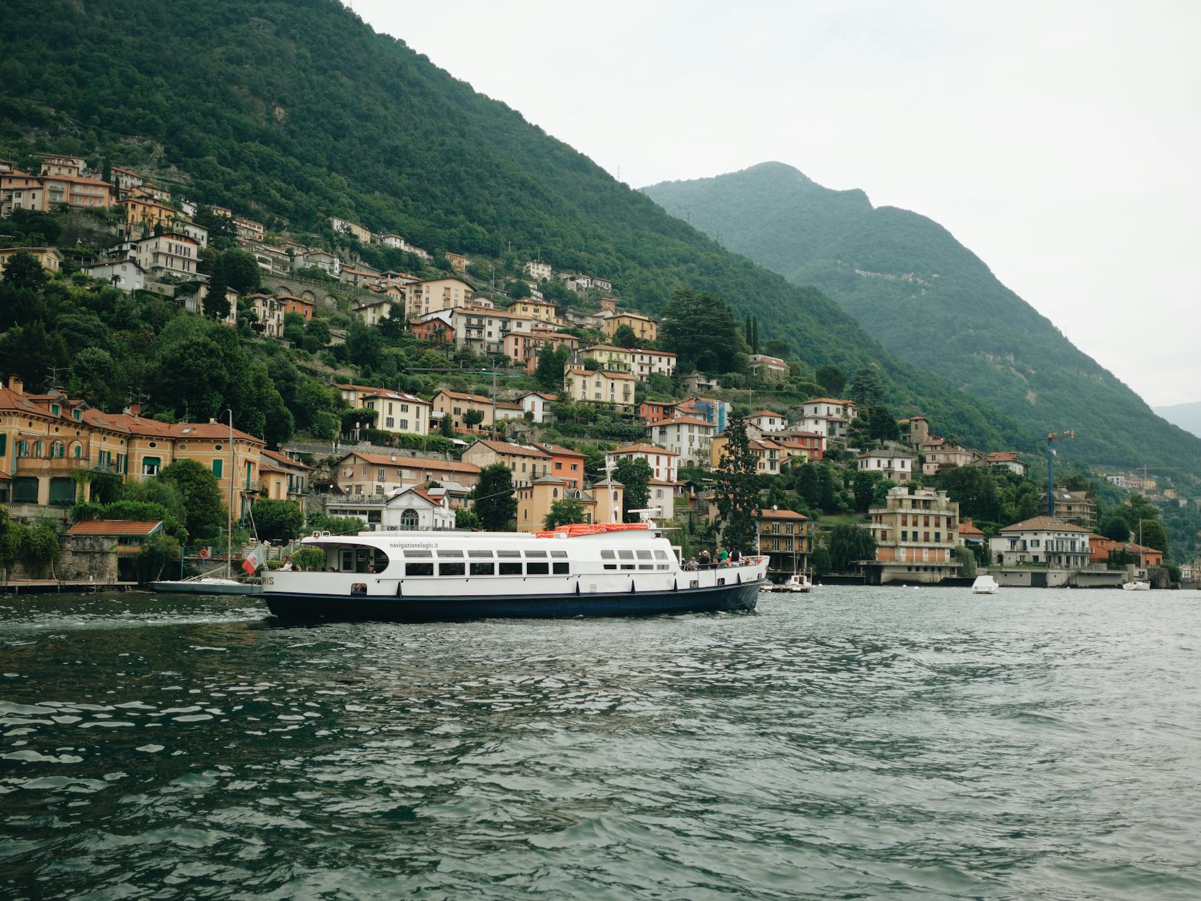 A ferry sails on Lake Como with a picturesque hillside town in Italy, showcasing vibrant architecture.