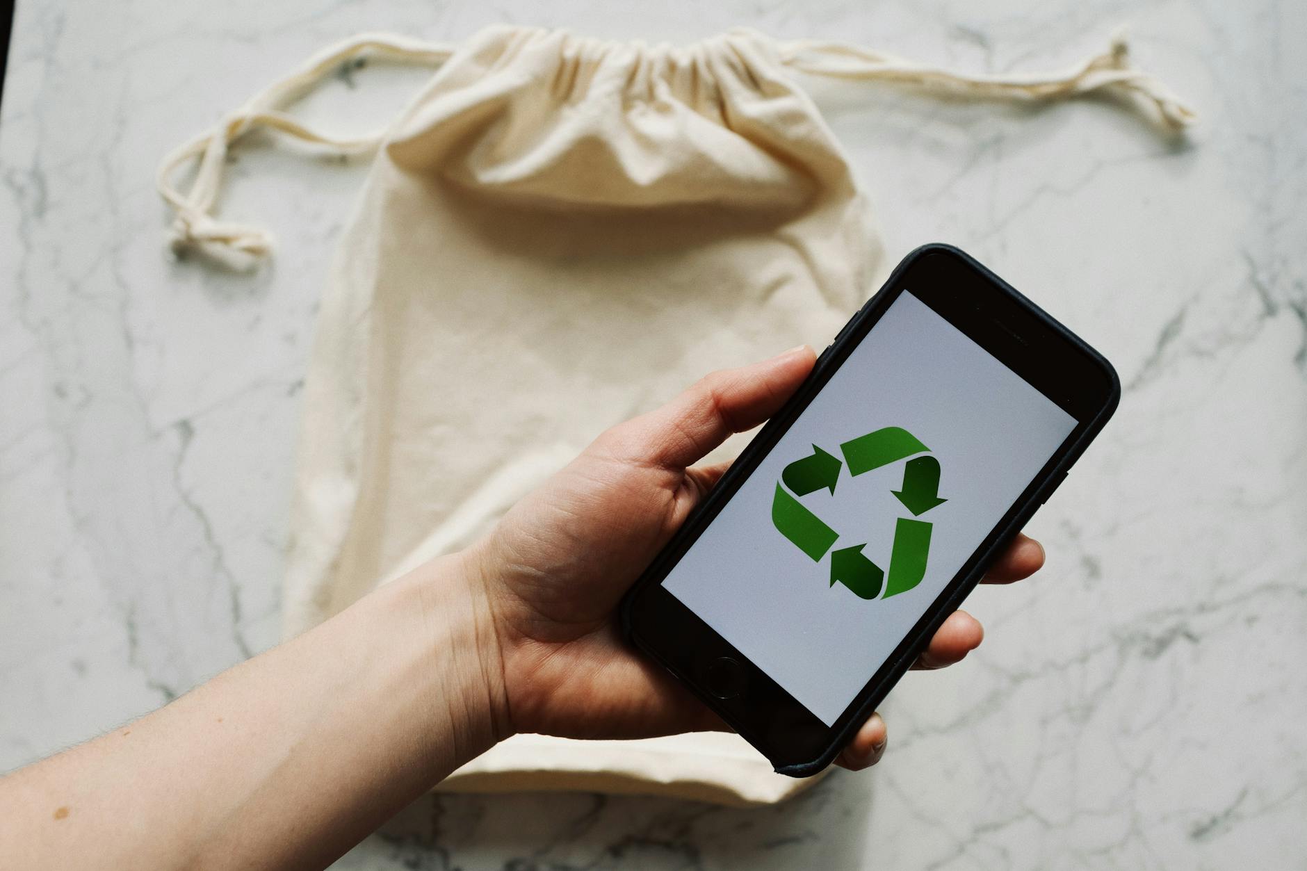 A hand holds a smartphone showing a recycling symbol over a reusable bag, promoting sustainability.