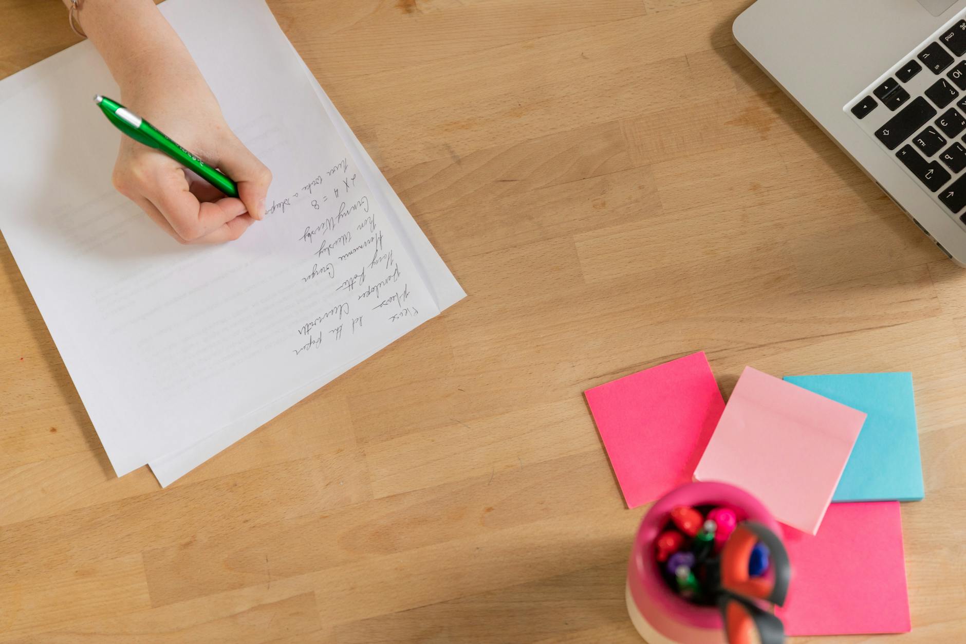A person's hand writing notes on paper next to a laptop and colorful sticky notes.
