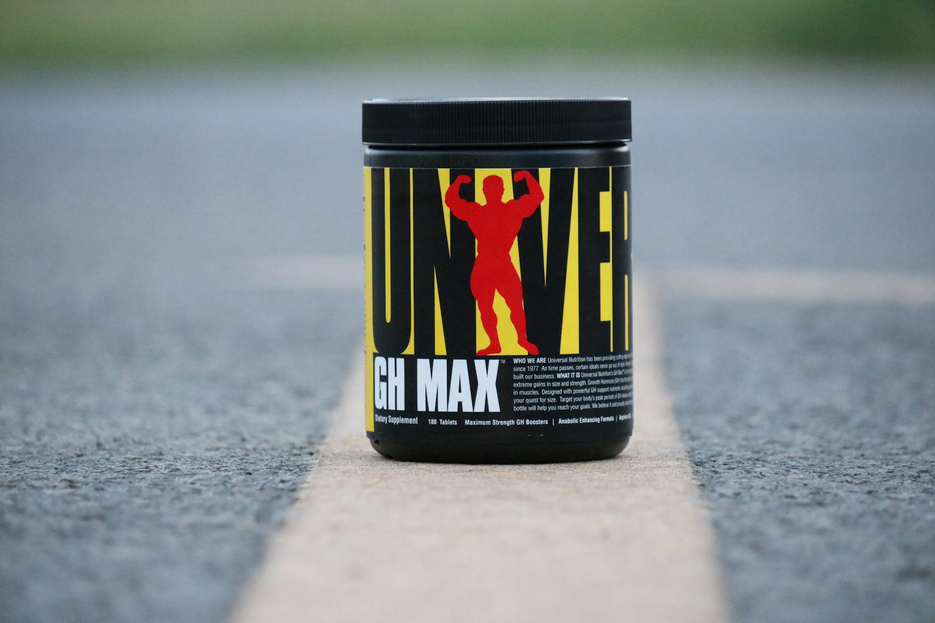 Black container of UNIVERSAL Nutrition GH MAX supplement on a road outdoors.
