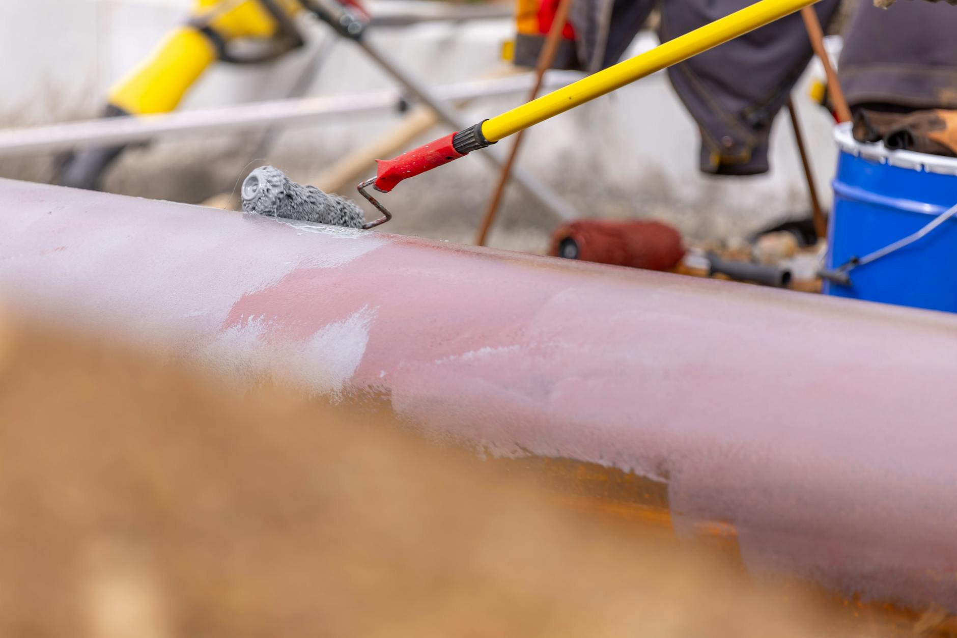 Close-up of a roller brush painting an industrial pipeline outdoors. Equipment visible in the background.