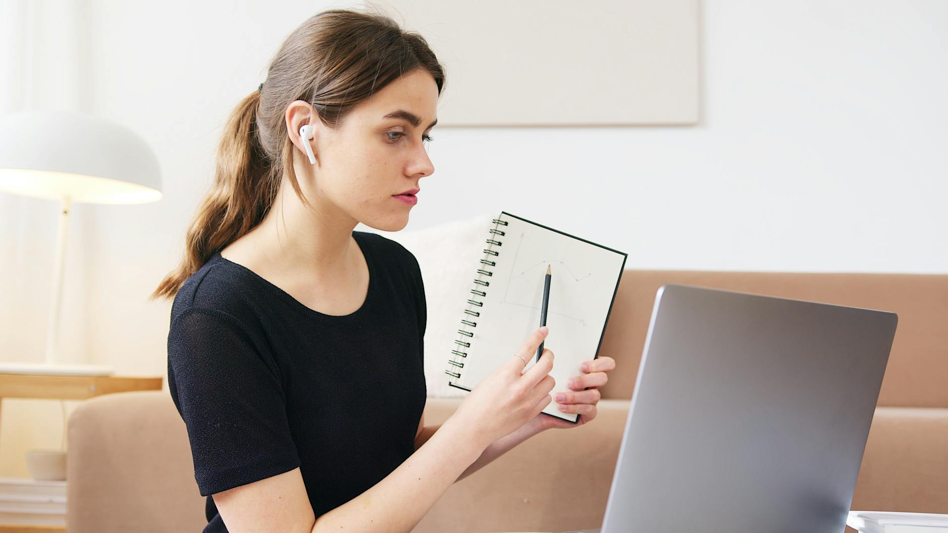 Serious young female student in casual black shirt participating in online webinar via laptop and showing notebook with homework during quarantine