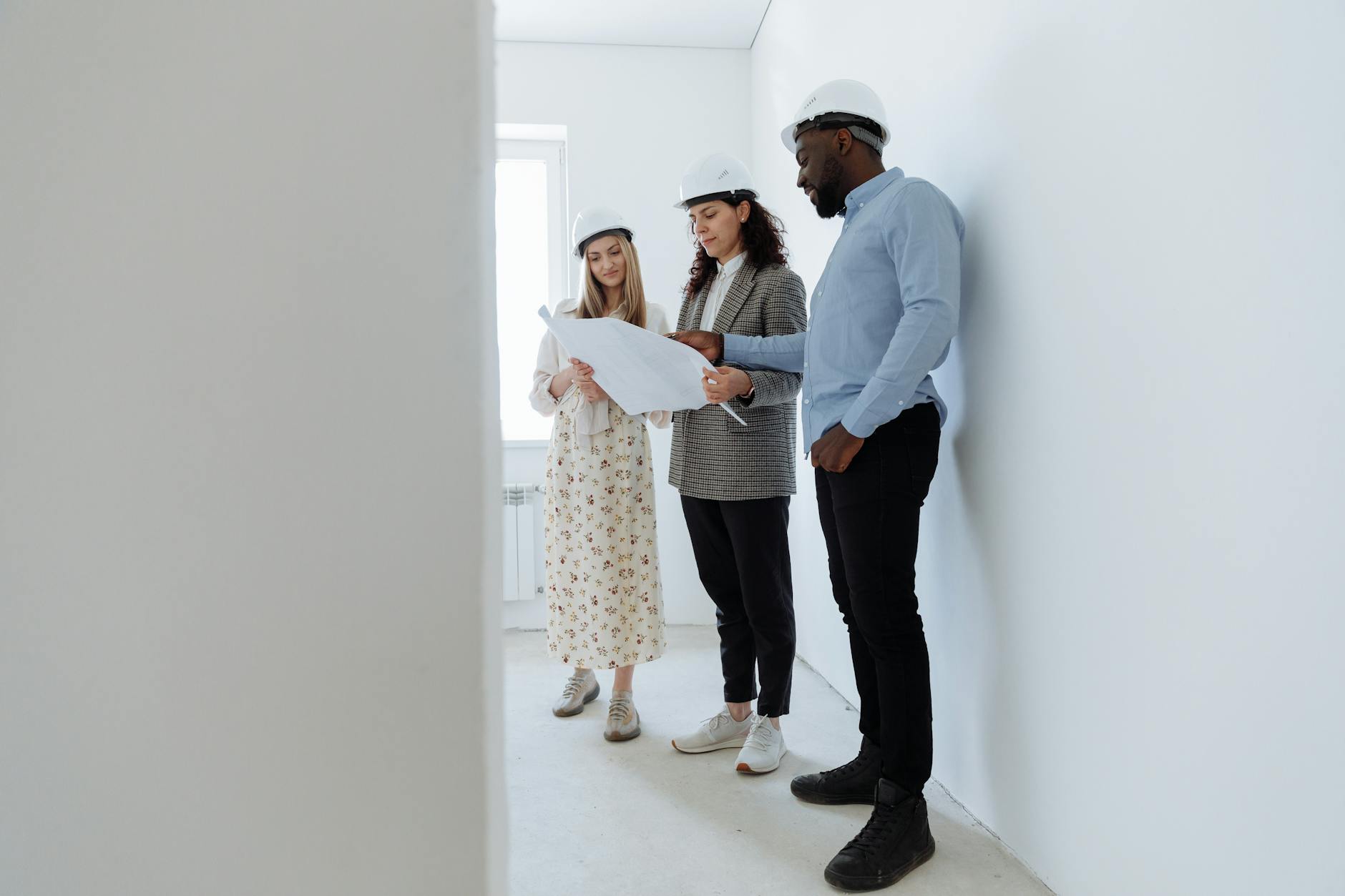 Three architects in hard hats review blueprints in a bright, empty white room.