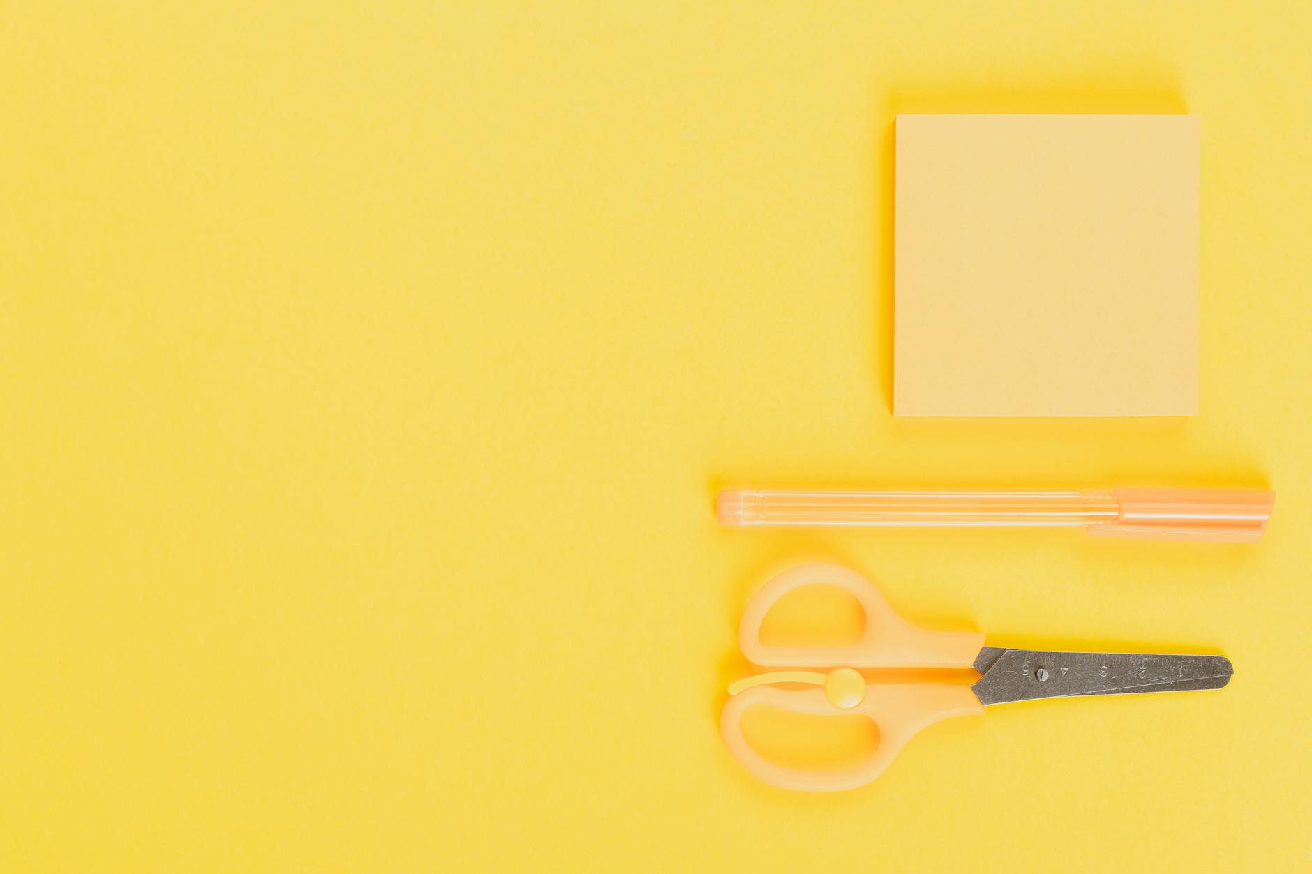 Top view of yellow school stationery including pen, scissors, and sticky note on yellow background.
