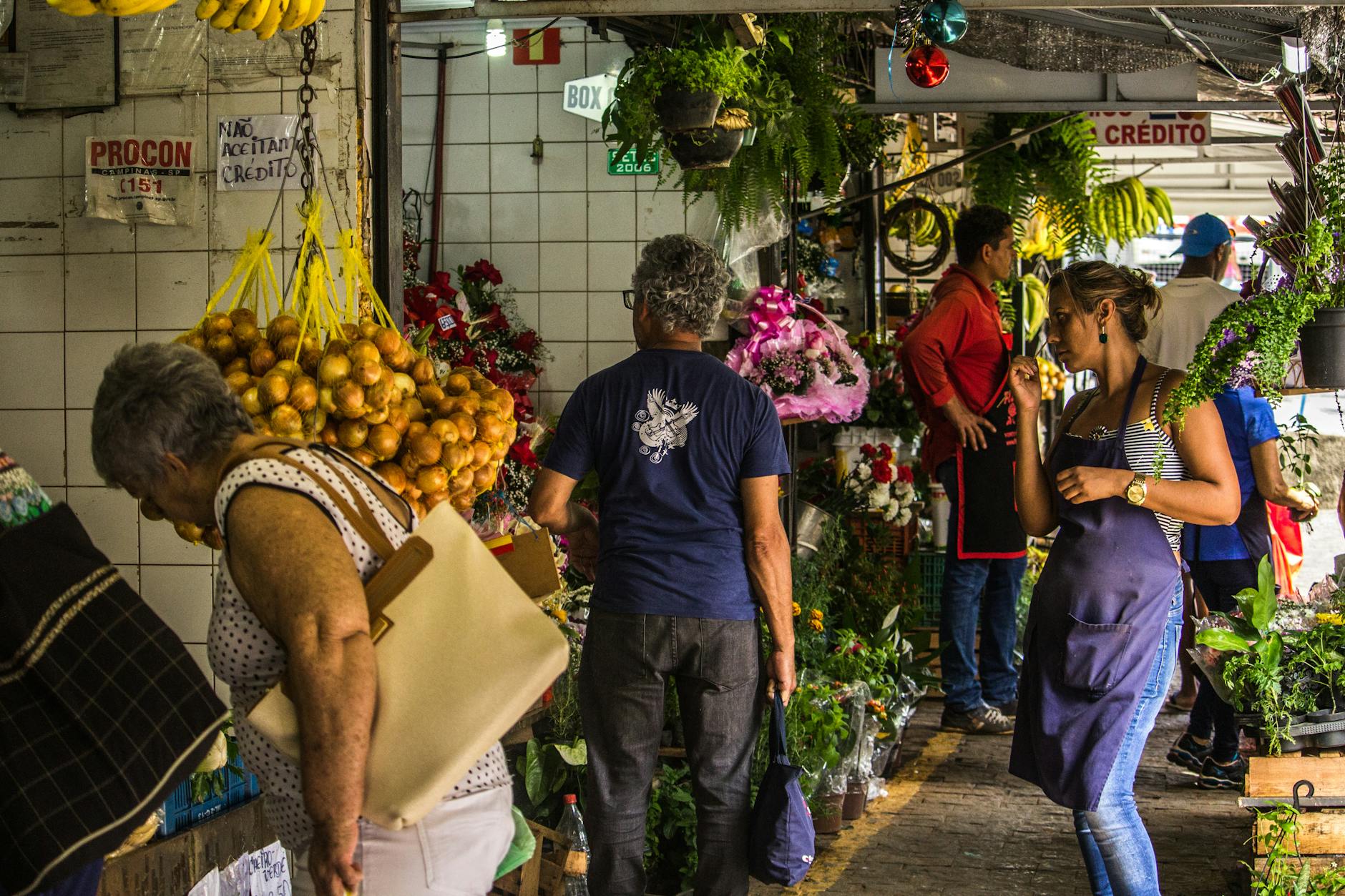 Vibrant indoor marketplace scene with diverse shoppers among colorful flower and fruit stalls.