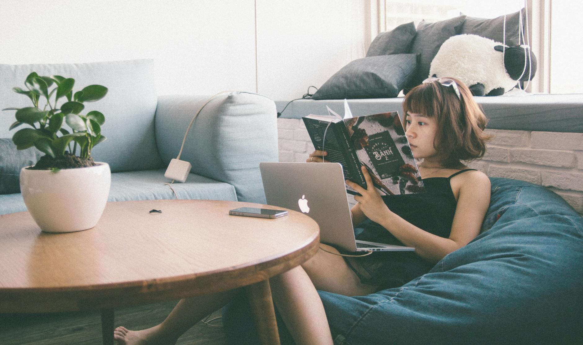 Woman reading while using a laptop in a cozy living space with modern decor.